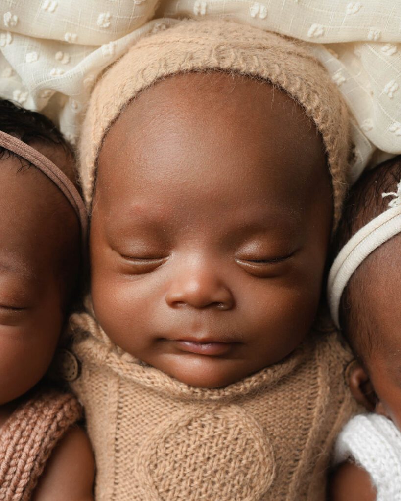Close-up of a newborn baby boy in a tan knit outfit and bonnet lying between his sisters.