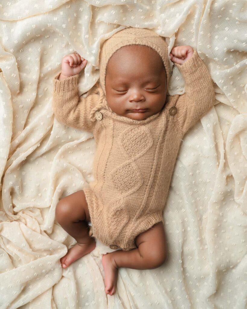 Sleeping newborn baby boy in a tan knit romper and matching bonnet with both arms raised.