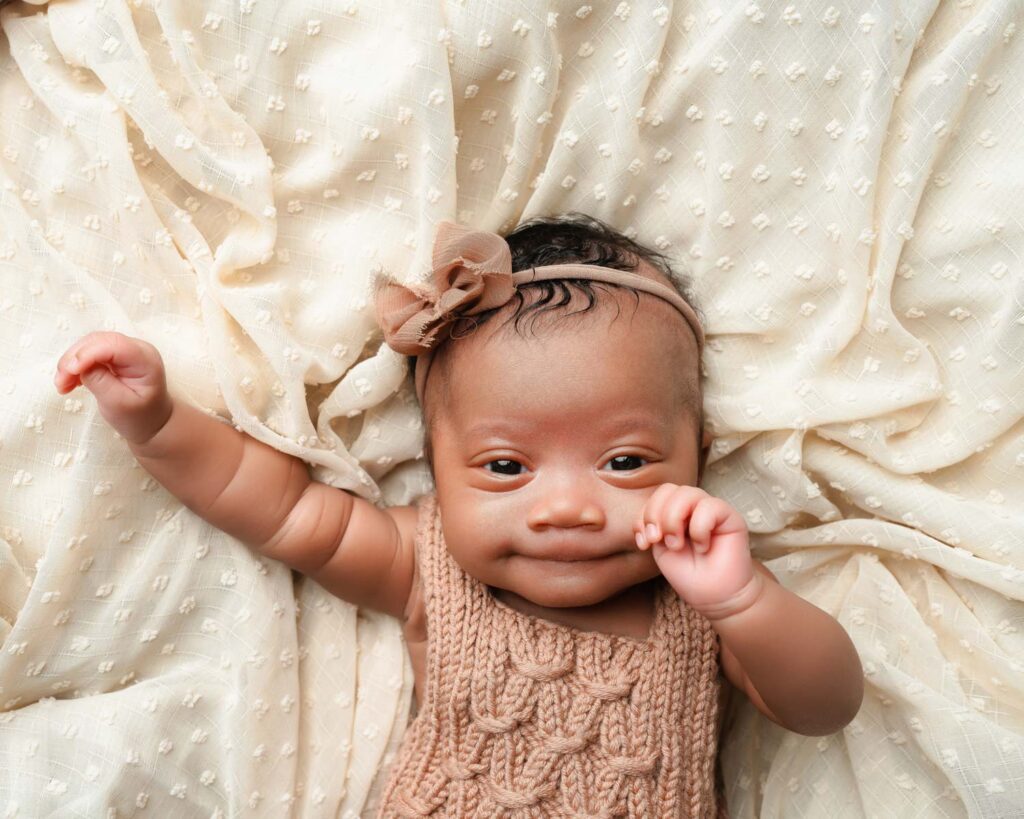 Awake newborn baby girl in a brown knit romper and bow headband resting on a cream blanket.