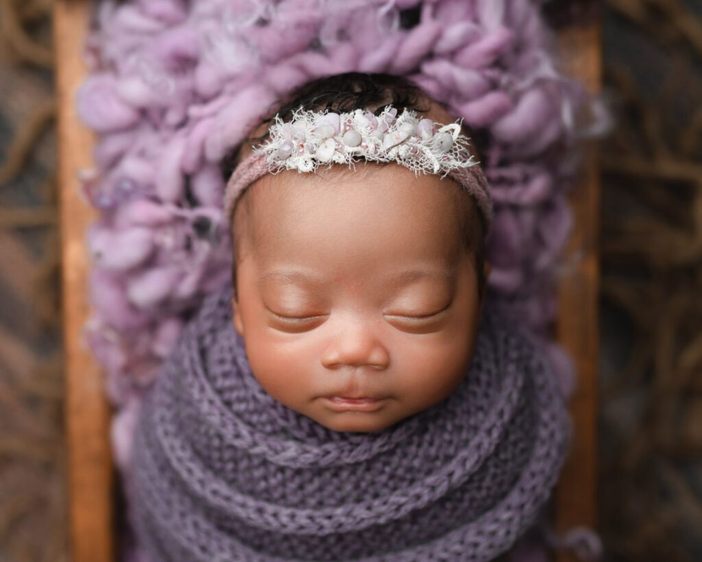 Sleeping baby girl wrapped in purple with a matching beaded headband on a soft purple blanket.