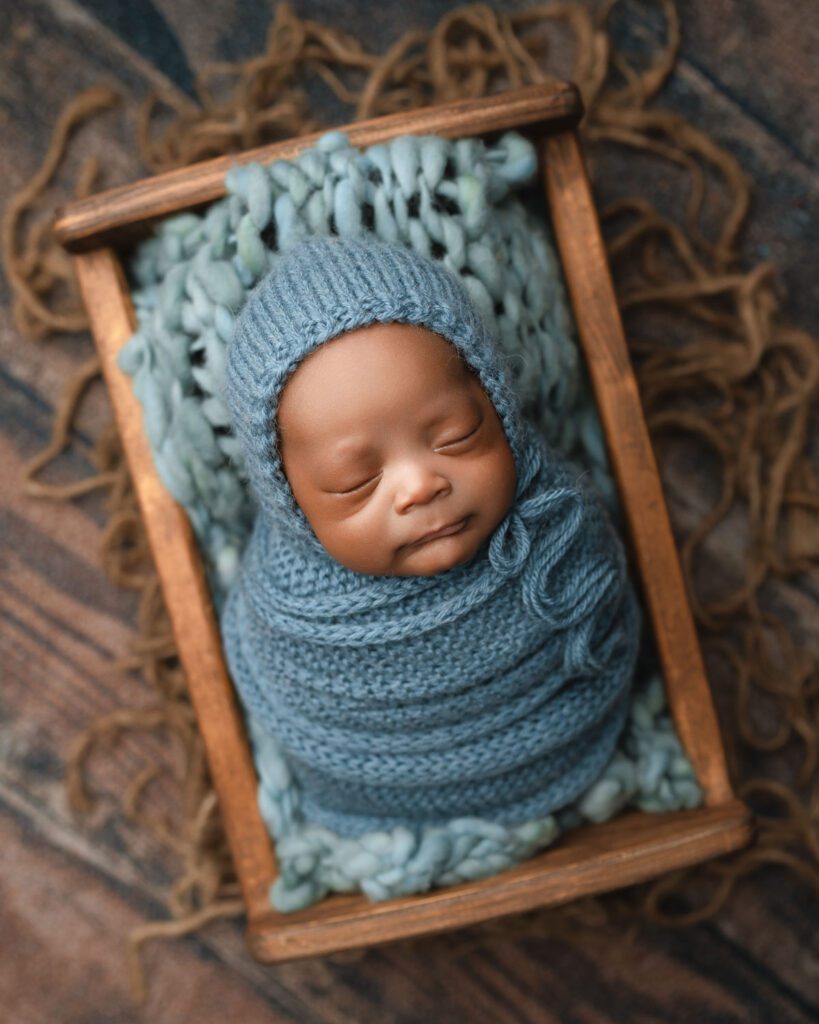 Newborn baby boy wrapped in blue crochet with a matching bonnet, resting in a wooden crate.