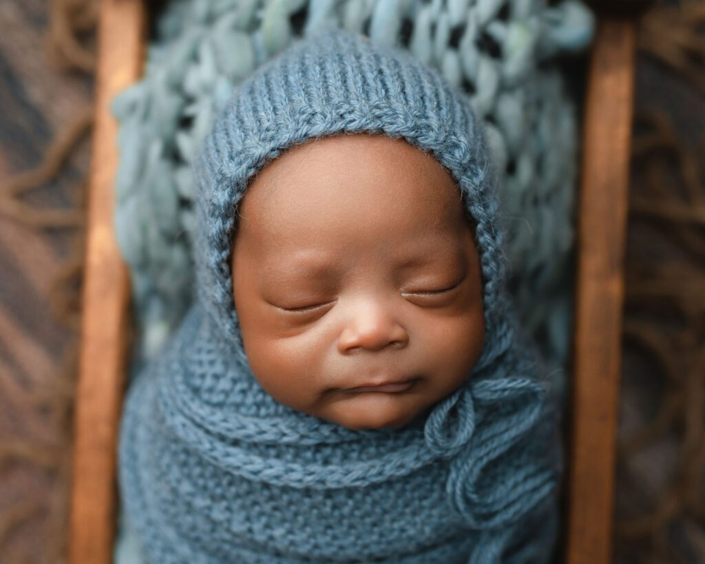 Sleeping newborn baby wrapped in blue knit fabric with a matching bonnet on a textured blue blanket.