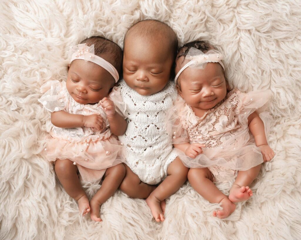Three newborn triplets resting together on a white fur blanket in coordinated white and blush outfits.