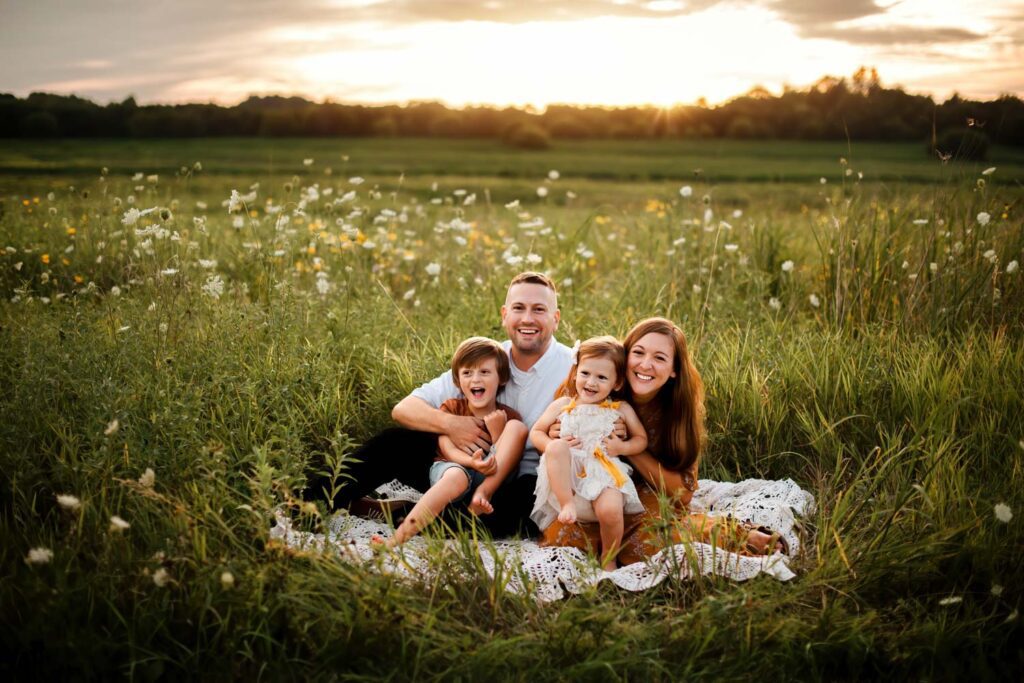 Mother and father snuggle on a blanket in a field of grass while holding their two giggling children at sunset at night.