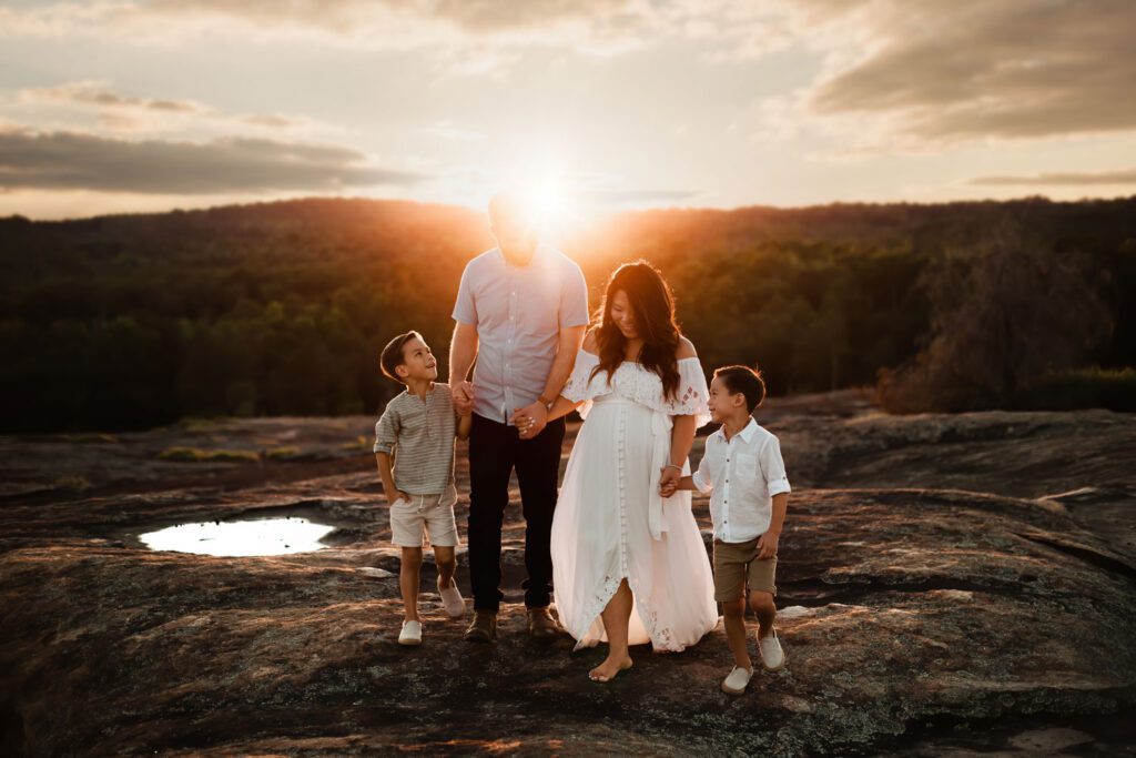 Pregnant mother and father hold hands with their twin boys and walk on a mountaintop at sunset with the sun shining.