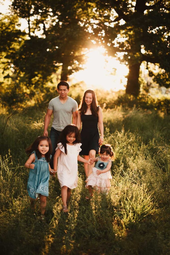 Family walking through grass as three daughters hold hands and walk in front of their parents in the golden hour evening.