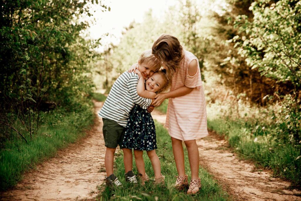 Three siblings hugging together on a narrow dirt trail bordered by tall grass and trees.