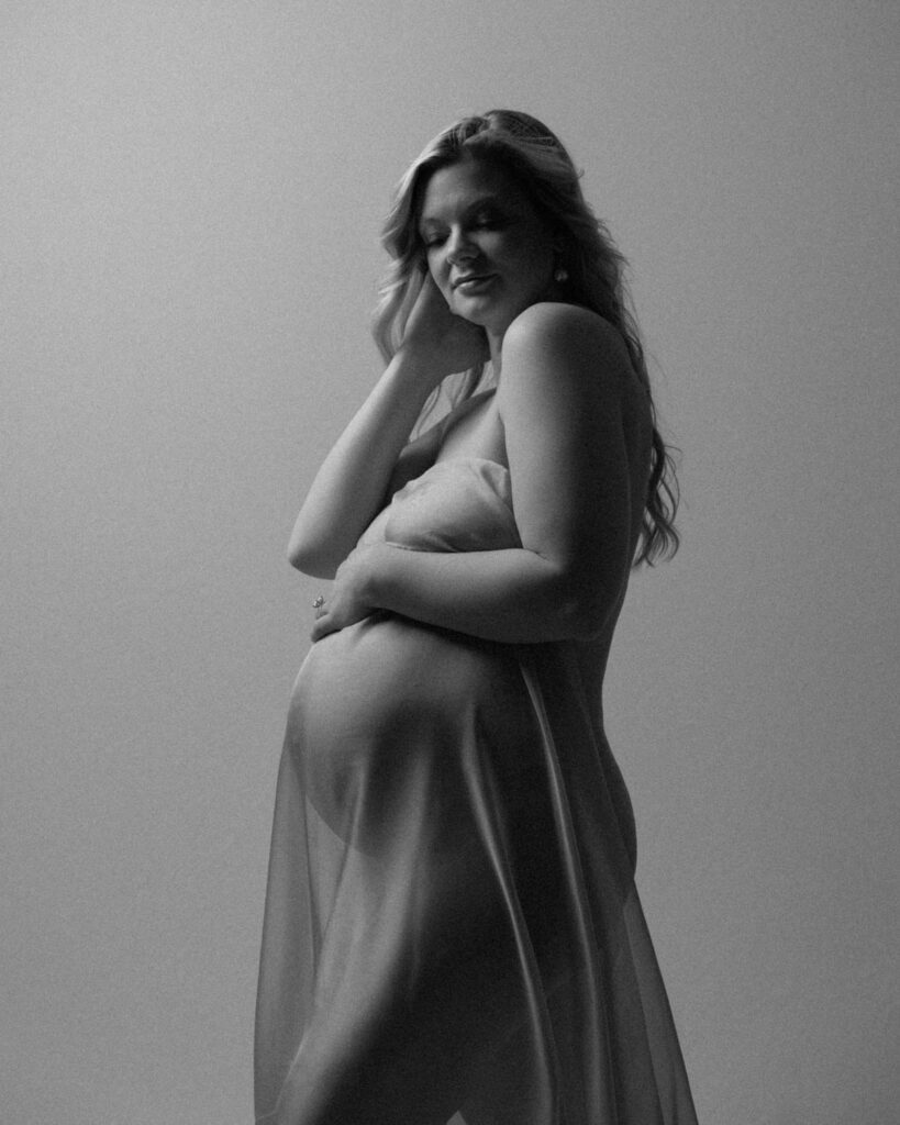 Black and white maternity portrait of an expecting mother in sheer fabric with one hand resting on her belly.