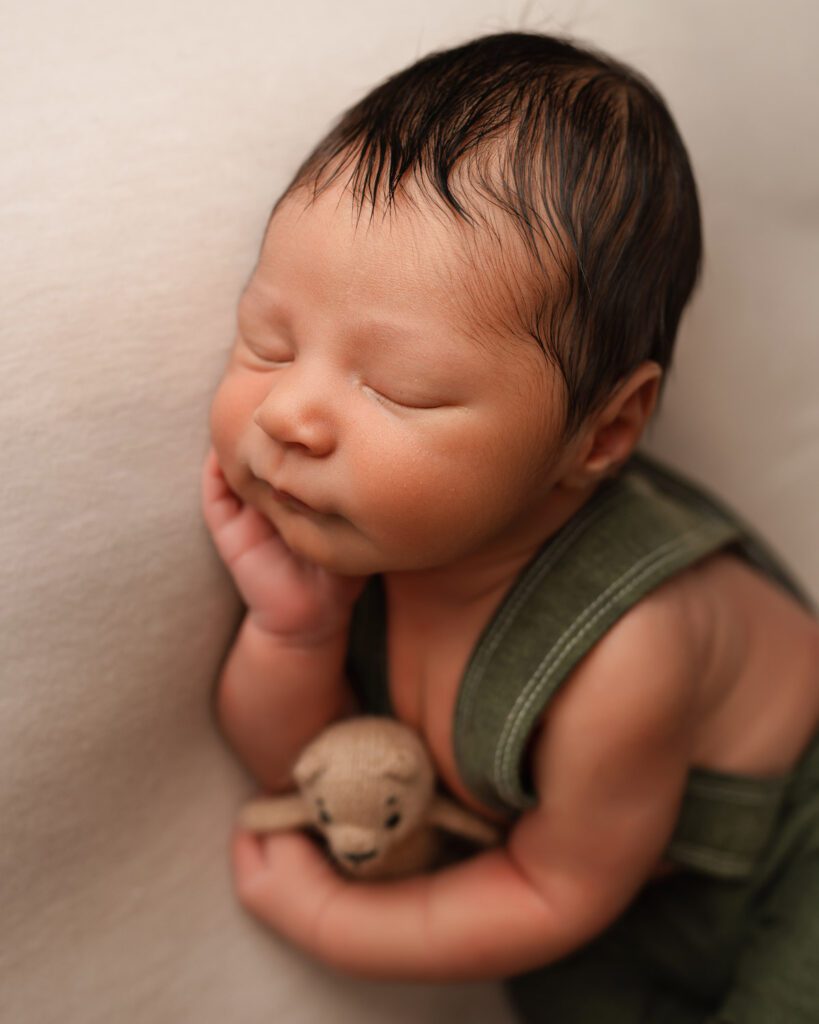 Close-up of a sleeping newborn in a green romper resting one hand on the cheek while holding a tiny bear.