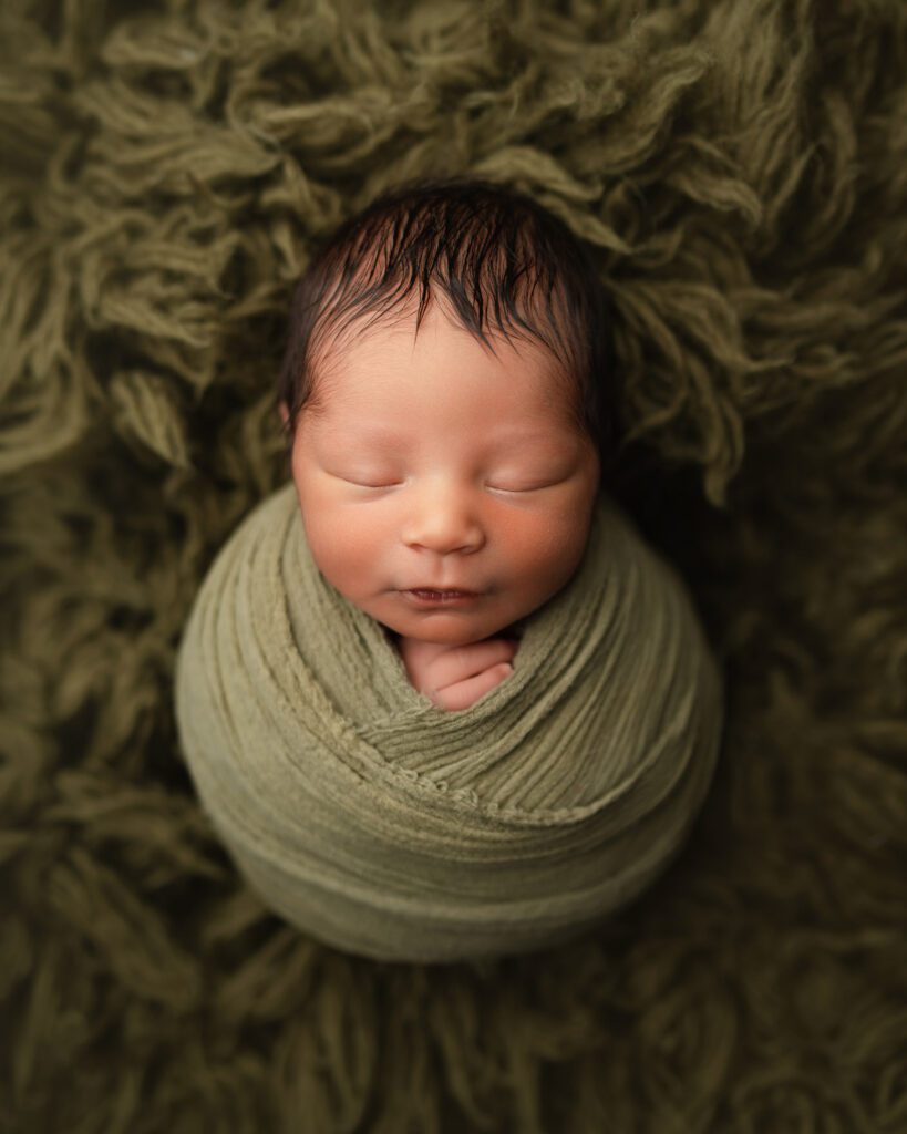 Newborn baby wrapped snugly in sage green and sleeping against a textured olive blanket.