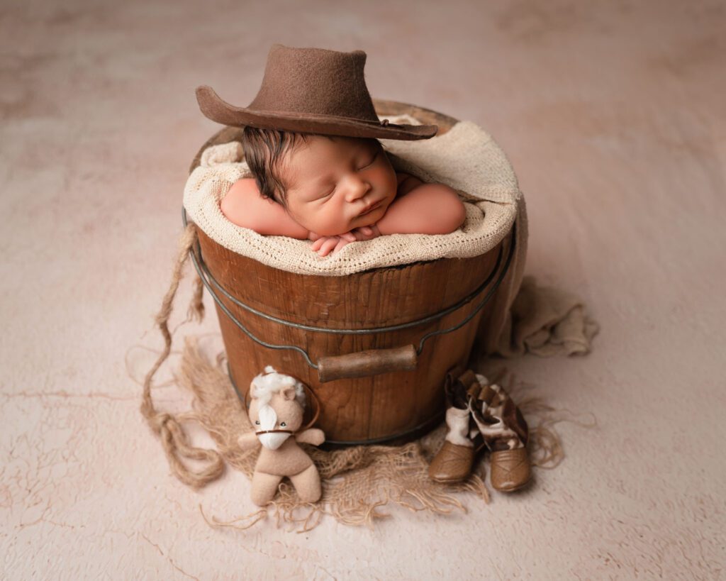 Sleeping newborn posed in a rustic wooden bucket with a cowboy hat and tiny boots and horse props.