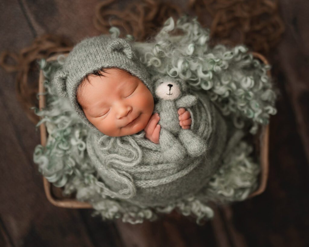 sleeping newborn boy wrapped in sage green knit wrap and teddy bear hat with a tiny stuffed bear.