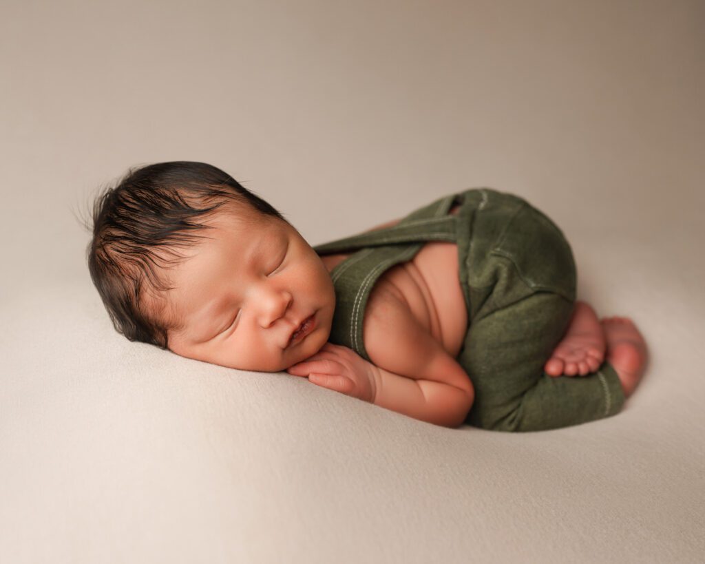 Sleeping newborn baby with dark hair wearing green pants and suspenders while posed on his stomach.