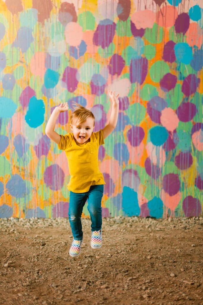 Young son in a yellow shirt jumping with both arms raised against a bright multicolored wall.