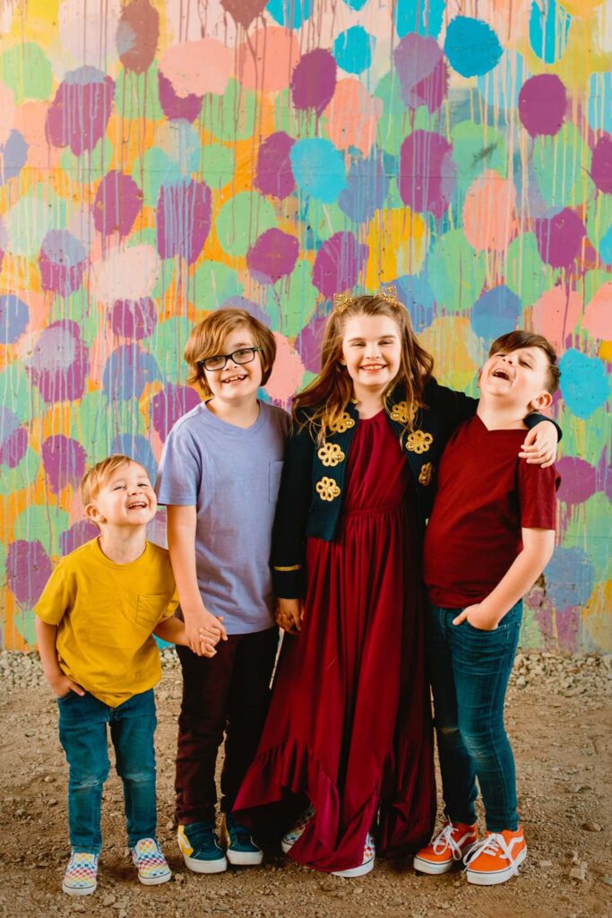 Four siblings laughing together in colorful outfits in front of a HENSE painted mural.
