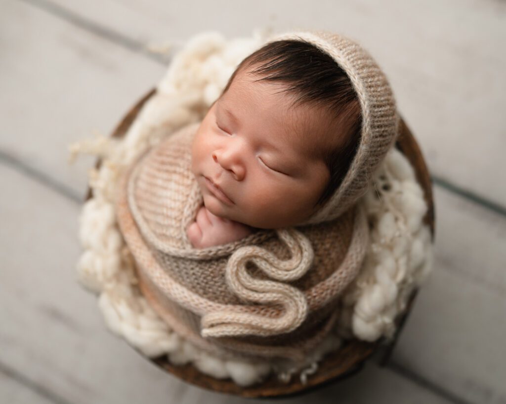 five week old newborn baby boy in tan swaddle wrap laying in bucket on top of white wooded floors.