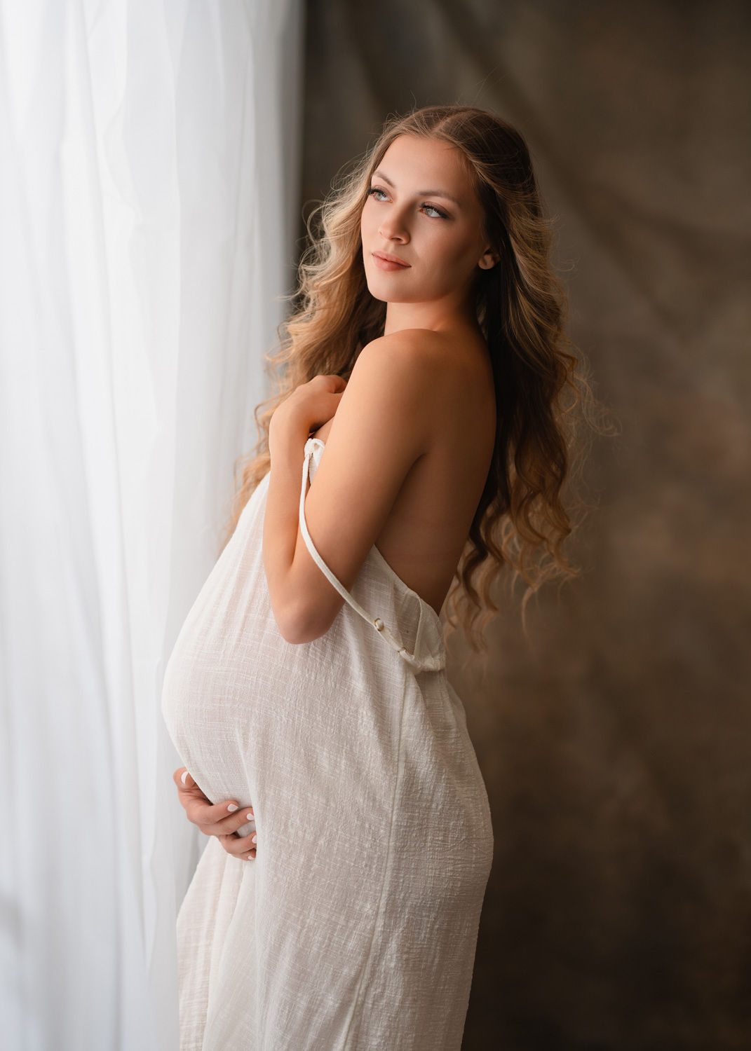 pregnant mom in sheer white dress looking out window in Atlanta, GA
