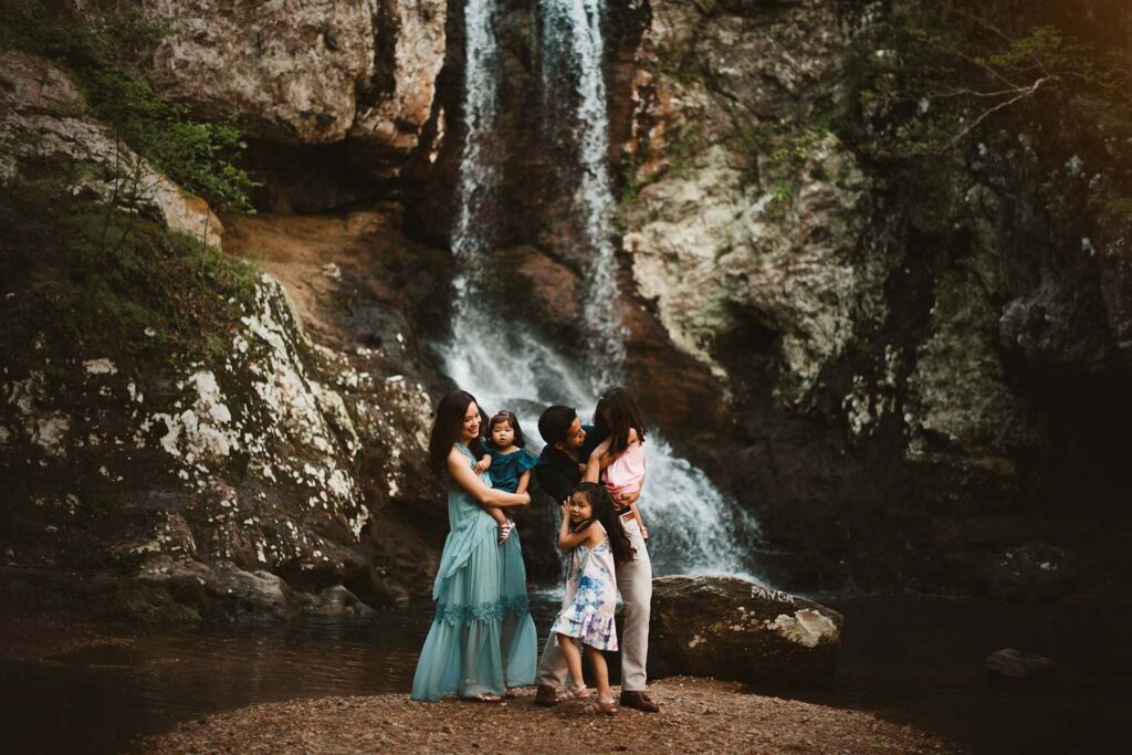Family of five wearing coordinating blue and pink clothes stand together in front of a waterfall on a tiny patch of land.
