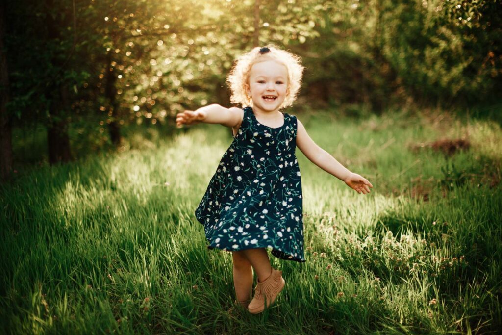 Happy little girl with blonde curly hair wears a blue dress with flowers spins around in the grass with her arms out wide. 
