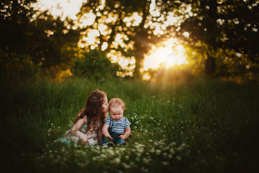 Siblings sitting together in a clover field at sunset as big sister with curly hair kisses her little brother on the head. 