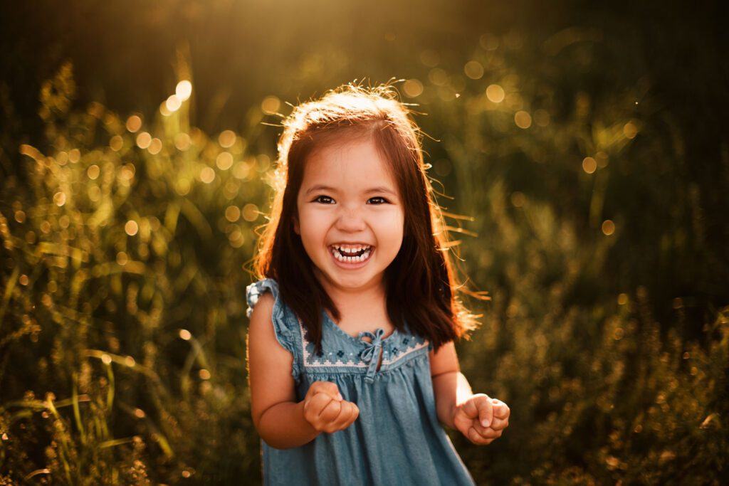 Little girl with dark brown hair wears a blue jean shirt stands in tall grass and smiles at the camera with sunlight behind. 