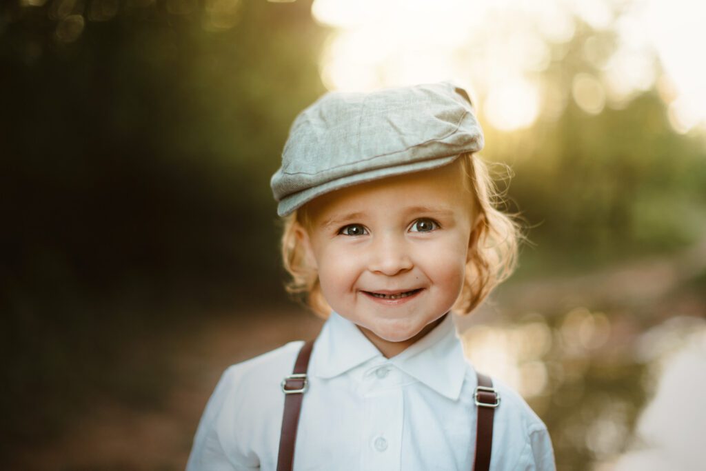 Blonde little boy wears a white button up shirt with brown leather suspenders and a grey newsboy cap hat smiles outside. 