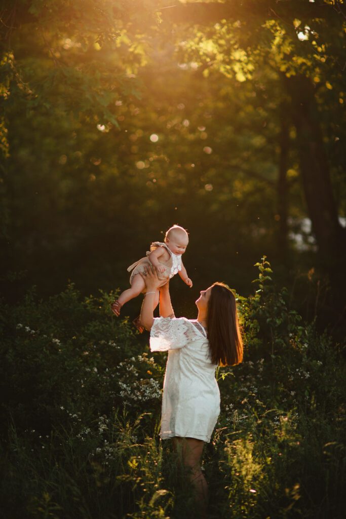 Mom in a white lace dress standing in tall grass holds her baby girl wearing a tan and lace onesie in the golden sunlight.