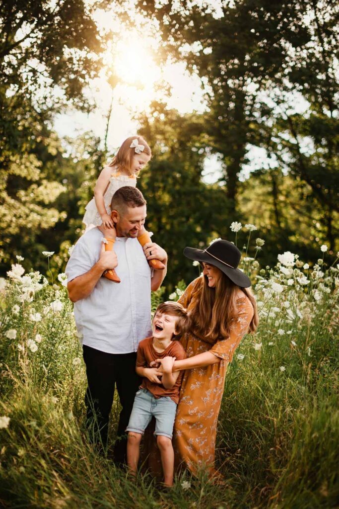 Laughing family standing in tall grass with white flowers as daughter is on dads shoulders and mom tickles her son. 