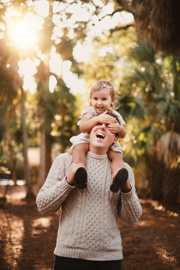 Little girl wearing a cream lace dress sits on her dads shoulders with her hands over his face as they both laugh at sunset. 