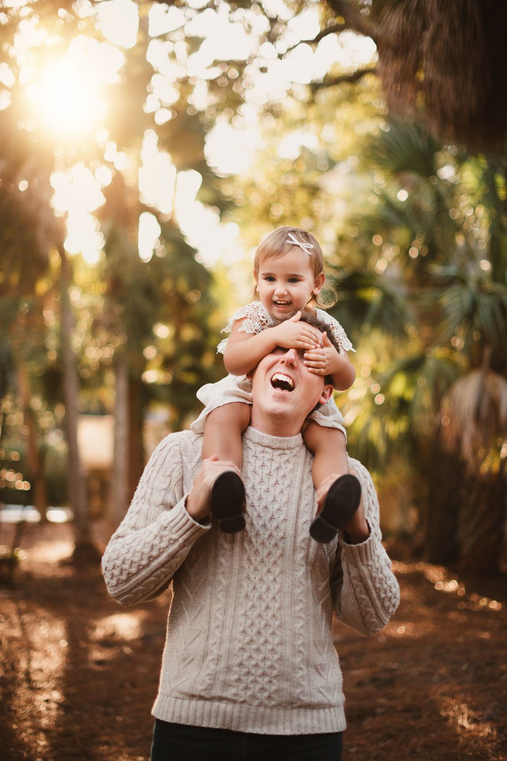 Little girl wearing a cream lace dress sits on her dads shoulders with her hands over his face as they both laugh at sunset.