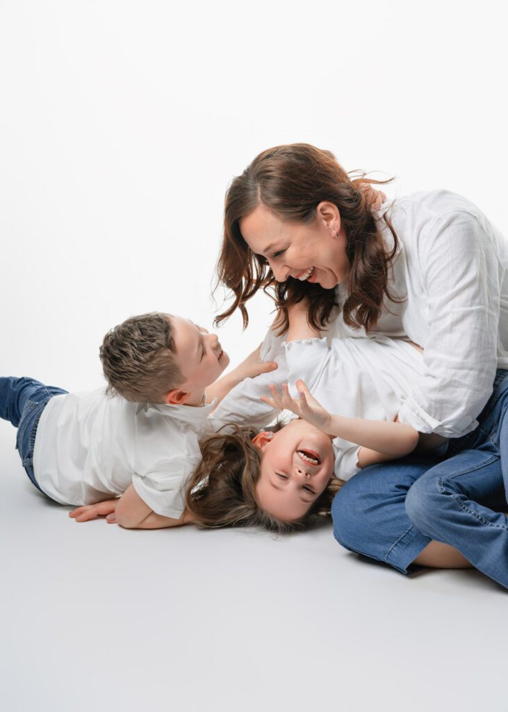 Mother tickles her son and daughter while wearing matching white shirts and blue jeans on a white backdrop in a photo studio.
