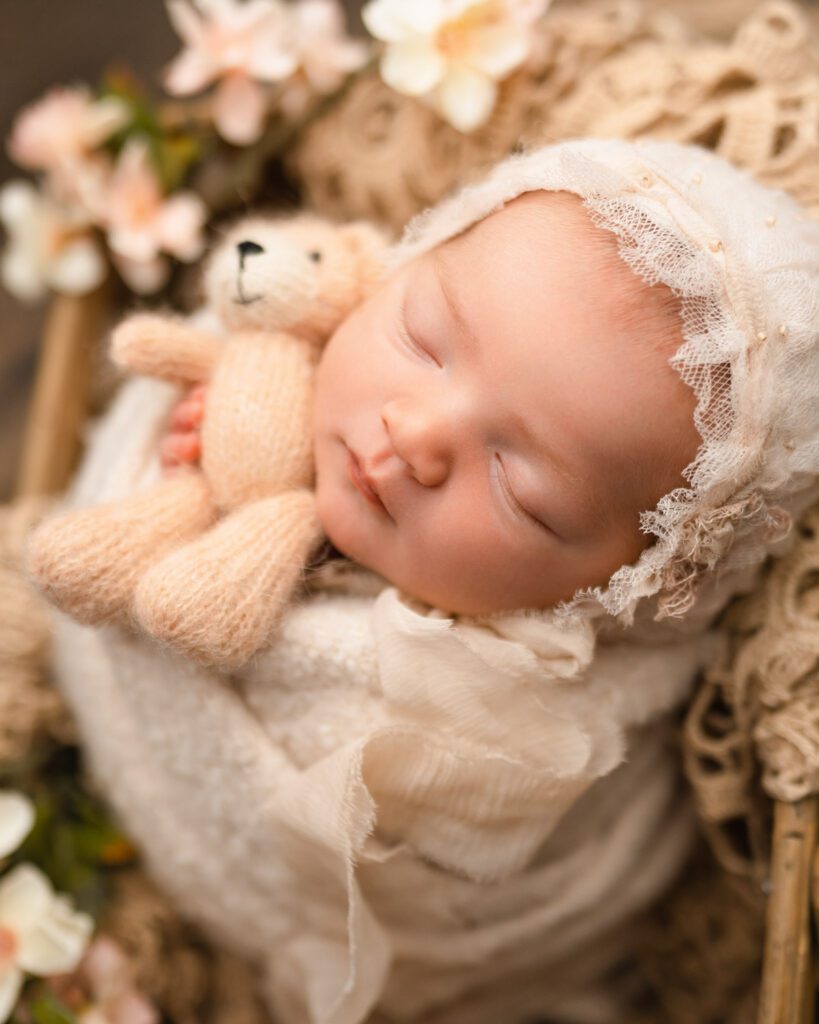 Dreaming newborn baby girl wears an antique bonnet holds a pink bear stuffy wrapped in a pink blanket surrounded by flowers.