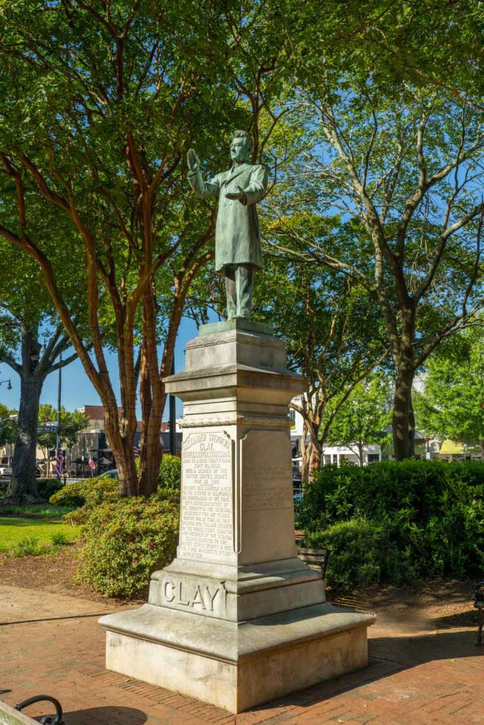Alexander Stephens Clay statue on a stone pedestal beneath leafy trees in Glover Park.