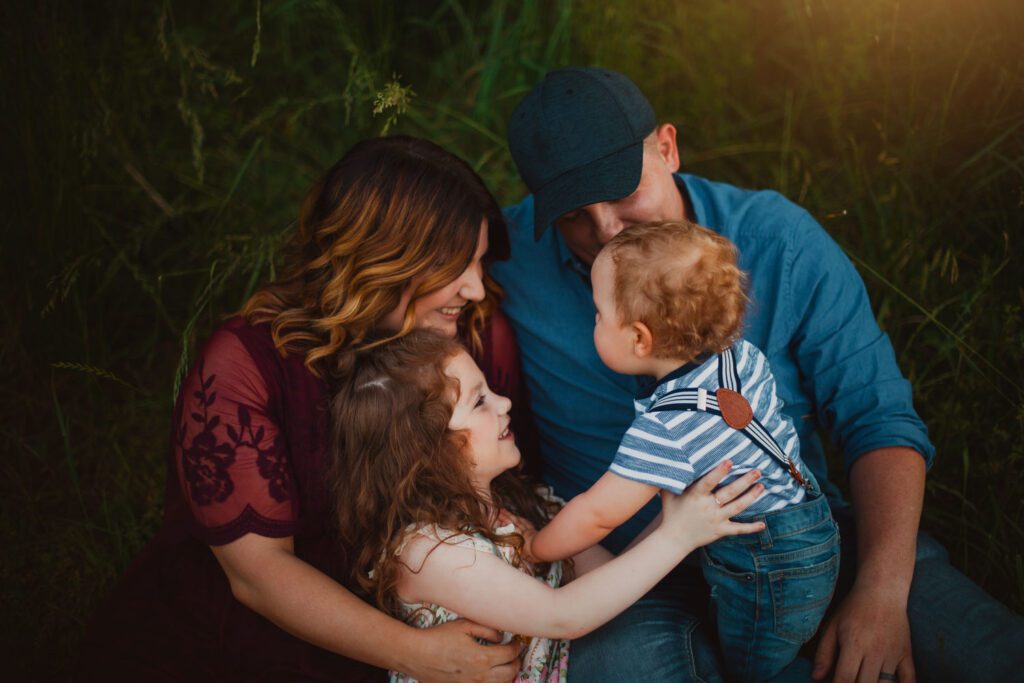 Parents cuddling their daughter and baby son together while seated in tall grass at sunset.
