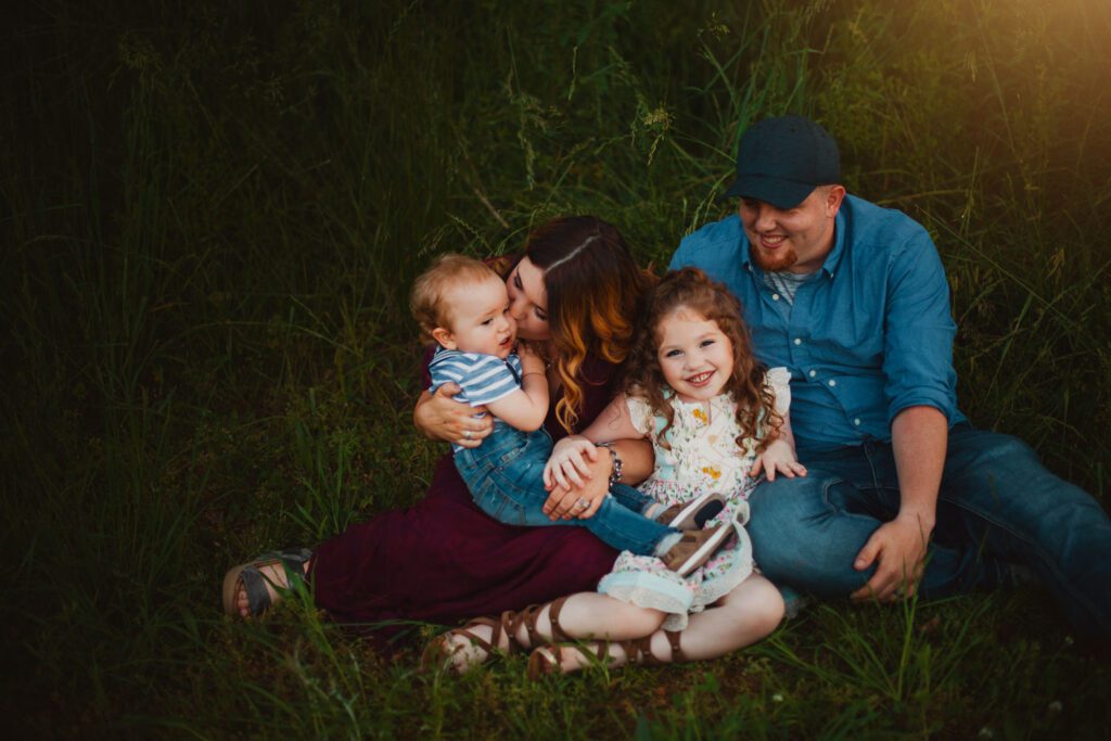 Mom and dad sitting in tall grass with their daughter and baby son gathered closely around them.