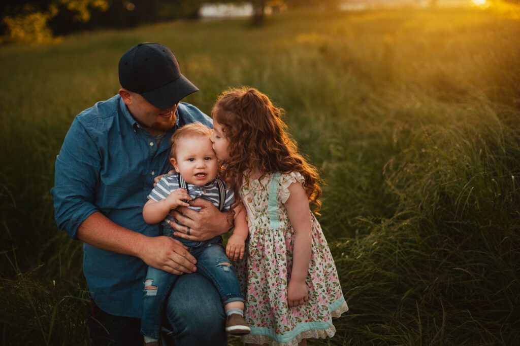 Dad holding his baby son while his daughter leans in to kiss the baby in warm evening light.
