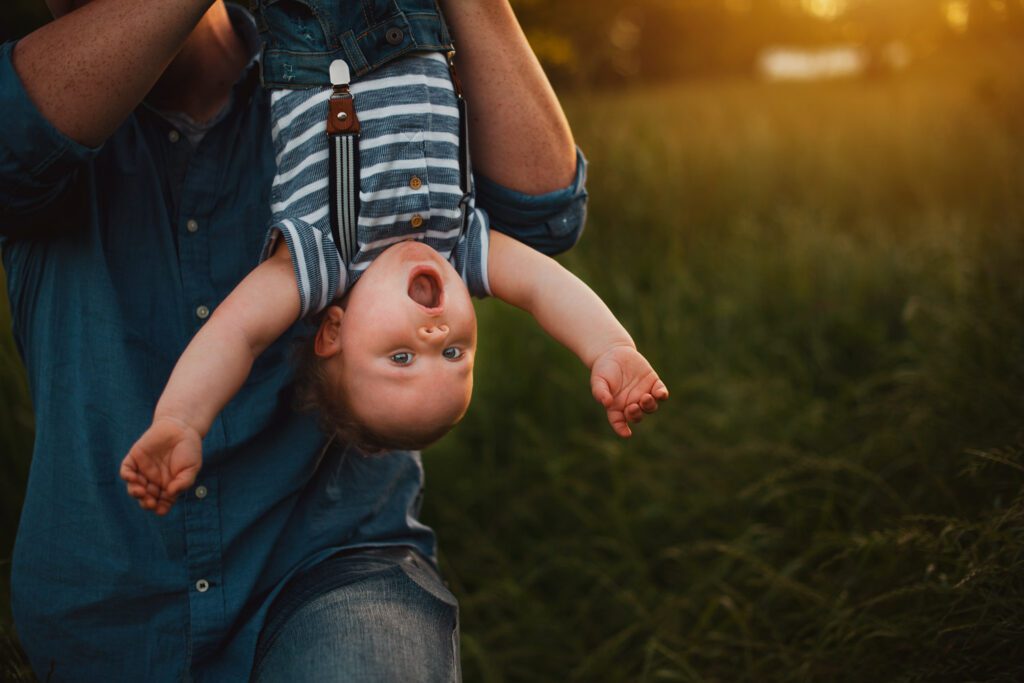 Dad holding his toddler son upside down by the legs as the boy looks surprised.