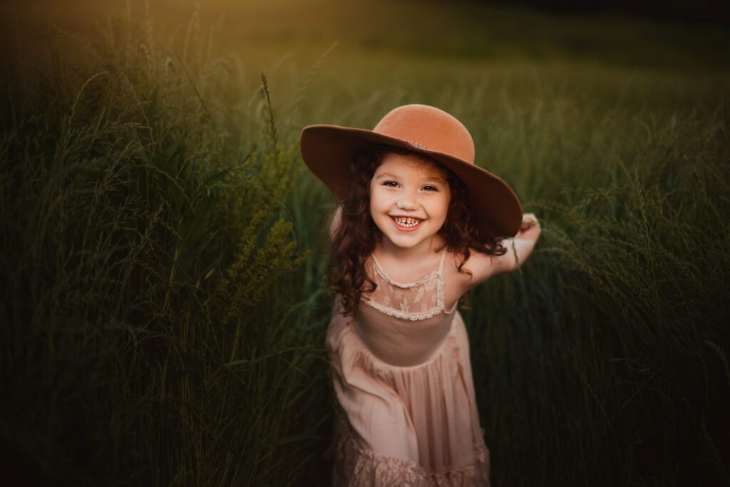 Young daughter in a blush dress and wide-brim hat leaning playfully toward the camera in tall grass.