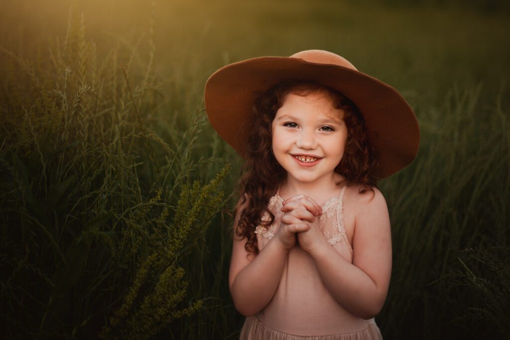 smiling young girl in wearing blush dress and brimmed hat clasping hands together in a meadow at sunset