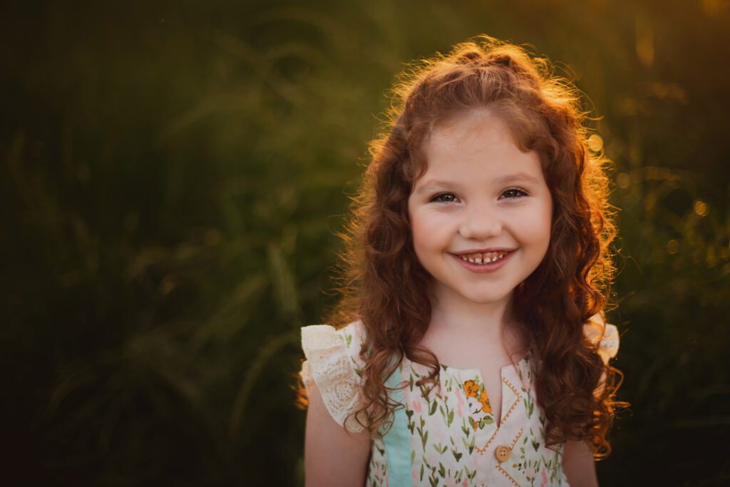 Close-up portrait of a smiling young girl with curly red hair and a floral dress in warm evening light.