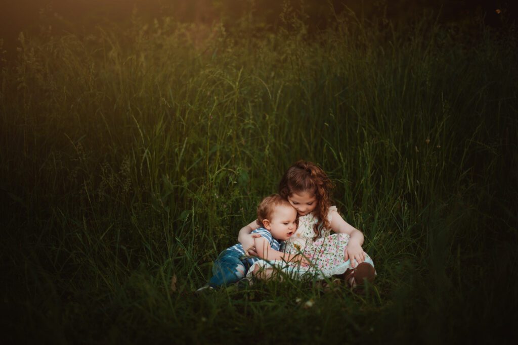 Older sister cuddling her baby brother while they sit together in tall grass at sunset.