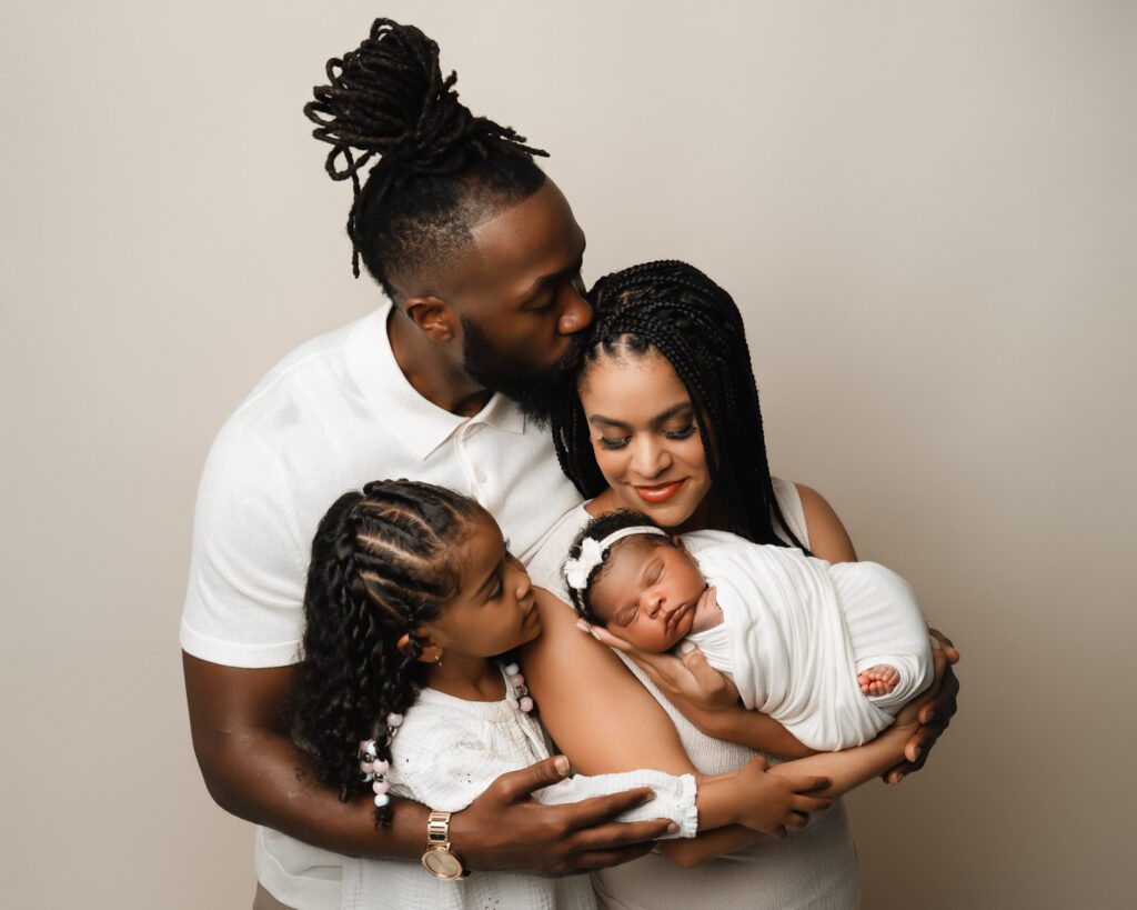 Family in white cuddling together against a neutral cream backdrop while looking at their newborn baby girl.