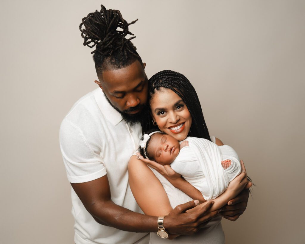 Mom and dad cuddling their swaddled newborn daughter together against a simple cream backdrop.