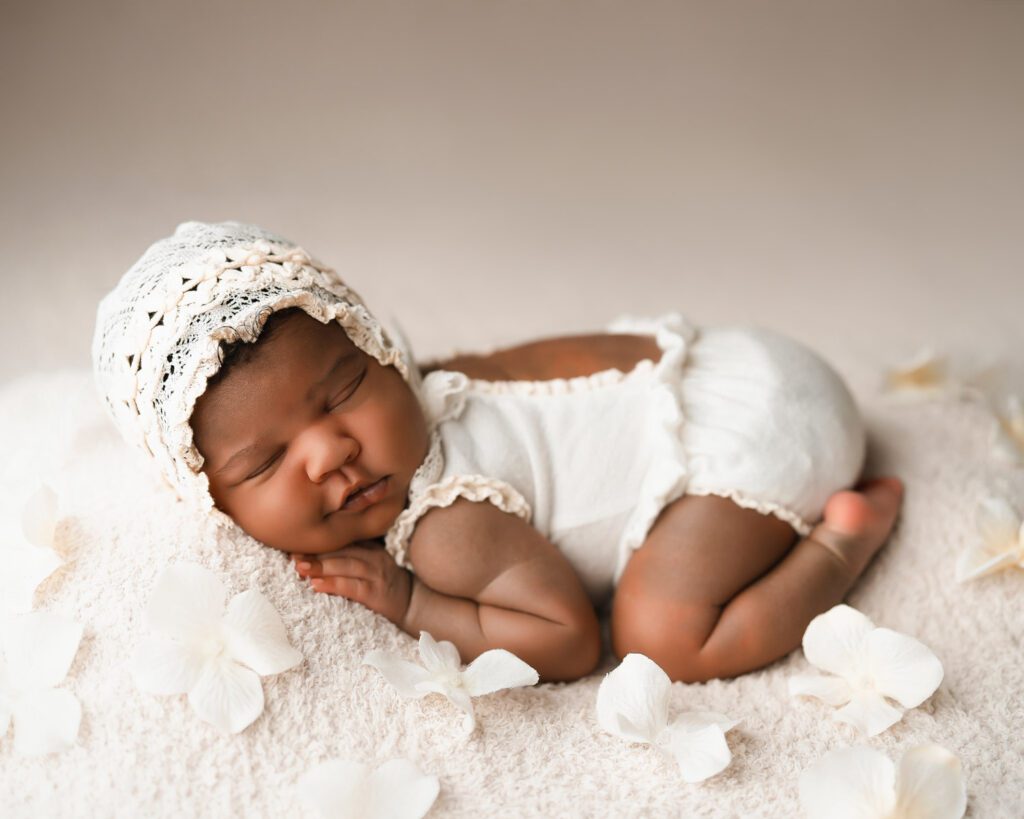 Sleeping newborn baby girl in a white onesie and lace bonnet surrounded by soft flowers on a plush blanket.