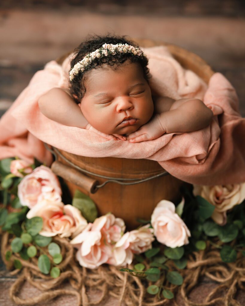 Newborn baby girl sleeping in a wooden bucket with a blush blanket, pink flowers, and greenery on a hardwood floor.
