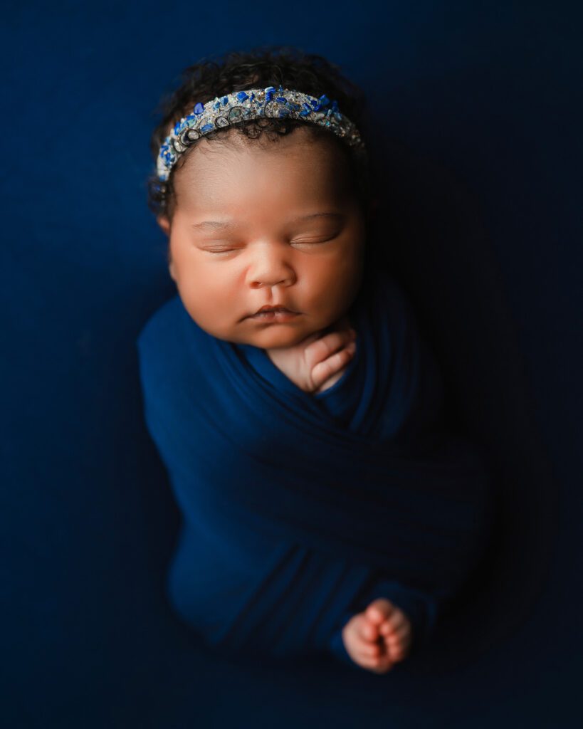Sleeping newborn wrapped in navy blue with a crystal headband on a matching blue fabric backdrop.