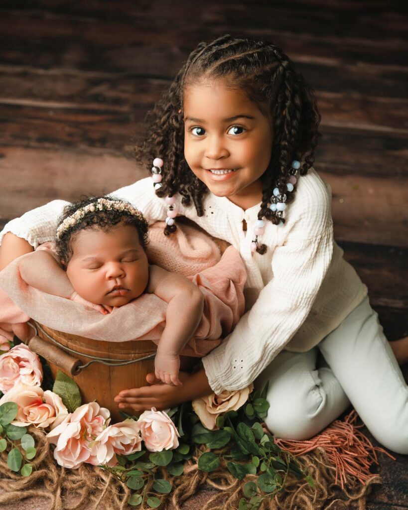 Big sister cradling a wooden bucket as her newborn sister sleeps inside surrounded by pink flowers.