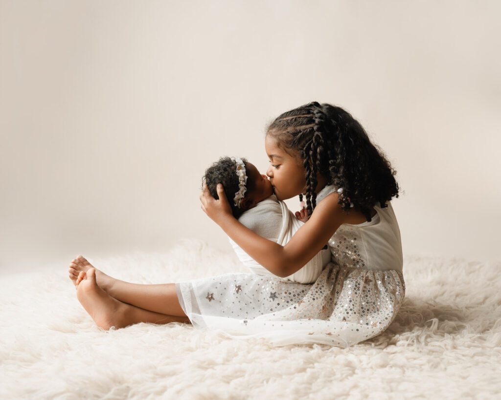 Big sister gently kissing her newborn sister while holding her on a soft cream rug.