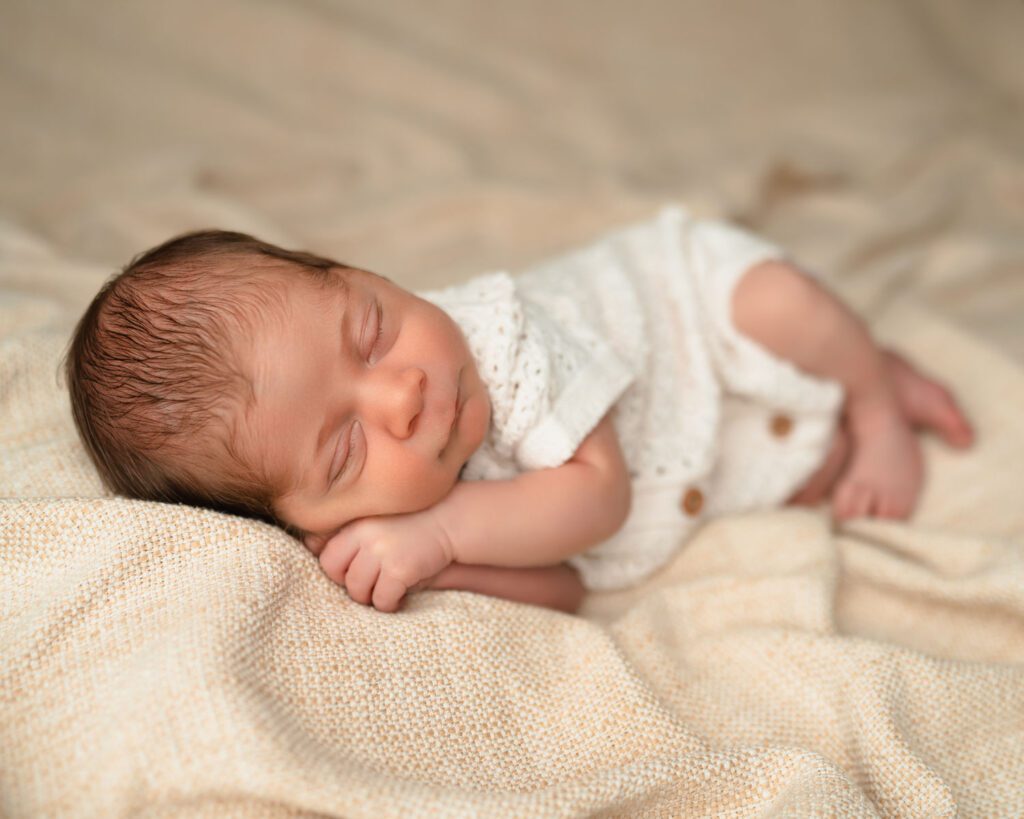 9 day old newborn baby boy laying on his side with hands tucked under his face wearing knit romper.
