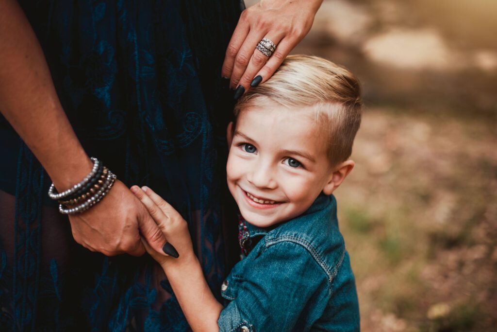 Smiling young son in a denim shirt holding mom's hand against a softly blurred woodland background.